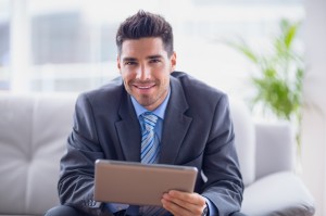 Businessman sitting on sofa using his tablet smiling at camera